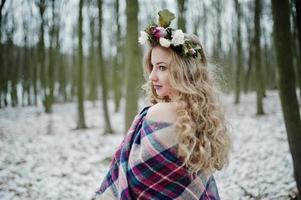 krullend schattig blond meisje met krans in geruite plaid op besneeuwde bos in winterdag. foto