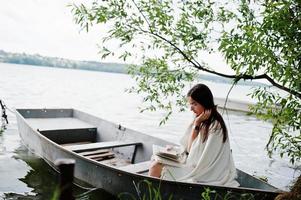 portret van een aantrekkelijke vrouw met zwarte stippenjurk, witte sjaal en bril die een boek leest in een boot op een meer. foto