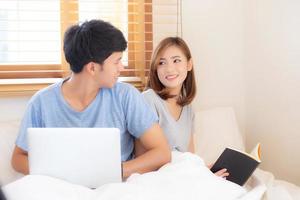 Aziatisch jong stel met man met laptopcomputer en zoeken op internet en vrouw die notitieboekje leest op bed in de slaapkamer met gelukkig en ontspannen, communicatie en bedrijfsconcept.