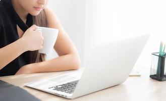 mooie jonge freelance aziatische vrouw die lacht werken en op laptopcomputer op bureau kantoor met professional, meisje met behulp van notebook en koffie drinken, business en lifestyle concept. foto