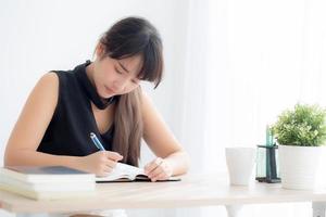 mooie jonge aziatische vrouw glimlachend zittend studeren en leren schrijven notebook en dagboek in de woonkamer thuis, meisje huiswerk, zakenvrouw werken aan tafel, onderwijs concept. foto