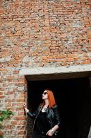 roodharige stijlvolle meisje in zonnebril dragen in het zwart, tegen verlaten plek met bakstenen muren. foto