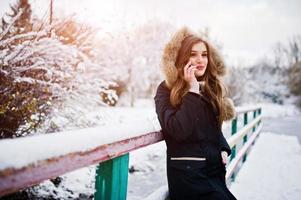 mooie brunette meisje in warme winterkleding. model op winterjas tegen bevroren meer in park spreken op mobiele telefoon. foto