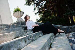 knappe jonge vrouw in witte blouse, wijde zwarte broek en zwarte klassieke hoge hakken zittend op trappen en poseren. foto