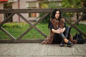 fashion outdoor foto van prachtige sensuele vrouw met donker haar in elegante kleding en luxe bontjas zittend tegen houten balustrades.