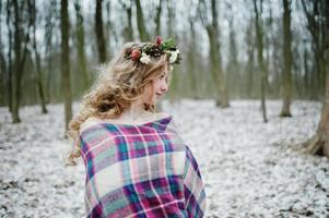 krullend schattig blond meisje met krans in geruite plaid op besneeuwde bos in winterdag. foto