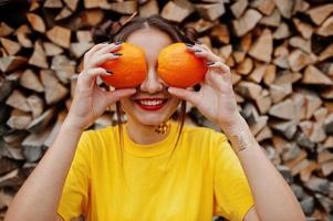 jong grappig meisje met lichte make-up, draag op geel shirt, houd twee oranje in de ogen tegen houten achtergrond. foto