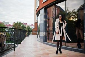 zwart haar sexy vrouw in bril en jas gesteld tegen gebouw met moderne ramen. foto