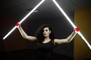 krullend brunette plus size model met twee lange led-lampen op zwarte kamer. foto