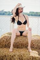 portret van een prachtig meisje in zwarte bikini zwembroek poseren op de hooibaal met een hoed aan het meer. foto