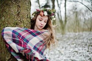 krullend schattig blond meisje met krans in geruite plaid op besneeuwde bos in winterdag. foto