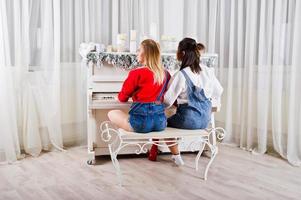 twee leuke, mooie meisjesvrienden dragen in overalls, spijkerbroeken, korte broeken en beenkappen tegen piano met nieuwjaarsversiering. foto