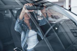 smartphone liggen. schattig meisje met zwart haar probeert haar gloednieuwe dure auto in de autosalon foto