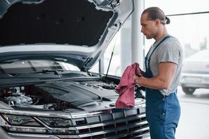 handdoek in handen. man in blauw uniform werkt met kapotte auto. reparaties uitvoeren foto