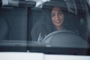 schattig meisje met zwart haar probeert haar gloednieuwe dure auto in de autosalon foto