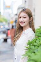 Aziatische mooie vrouw met lang bronzen haar draagt een wit shirt met lange mouwen en glimlacht gelukkig in een stedelijk buitenpark terwijl ze naar de camera kijkt. foto