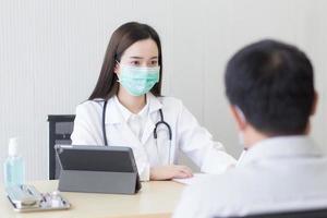 aziatische vrouw arts draagt een medisch gezichtsmasker en registreert symptoom van een man patiënt op papier in het ziekenhuis. coronavirusbescherming, nieuw normaal en gezondheidszorgconcept. foto