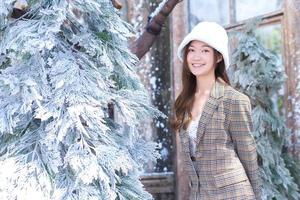 mooie aziatische vrouw draagt een witte hoed en geruite jas en staat in de buurt van met kerstboom bedekte sneeuw in nieuwjaars- en winterthema. foto