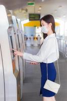 mooie jonge Aziatische zakenvrouw in een wit overhemd staat met een masker om ziektevirus en stof pm.2.5 te voorkomen om op een kaartautomaat in een luchttreinstation te drukken om op kantoor te gaan werken. foto