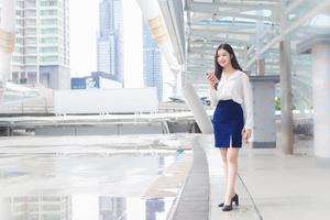 jonge aziatische zakenvrouw gaat naar kantoor of werkplek waar ze een smartphone in haar handen houdt in de grote stad met bedrijfsgebouwen met de stad als achtergrond. foto