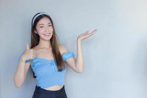 jonge mooie aziatische vrouw in een blauw shirt doet duim omhoog als een goed symbool en een andere hand toont iets op wit op de achtergrond. foto