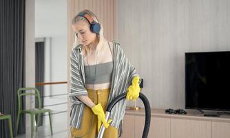 jong meisje heeft plezier tijdens het schoonmaken van de vloer met een stofzuiger. gelukkige vrouw die thuis huishoudelijk werk doet, geniet van muziek met oortelefoons. foto