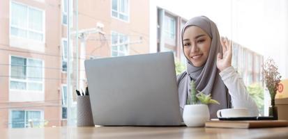 Glimlachende mooie moslim zakenvrouw met behulp van een laptop voor online videovergadering zittend aan de balie in moderne kantoorruimte, kijkt naar de webcam en zwaait hallo foto