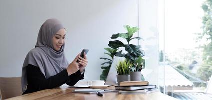 positieve mooie jonge moslimvrouw in paarse hoofddoek met behulp van gloednieuwe mobiele telefoon, kijkend naar kopieerruimte en glimlachen, met behulp van de nieuwste mobiele applicatie voor zaken, interieur foto