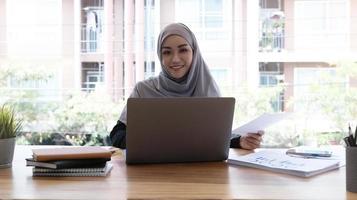 Aziatische moslim hijab vrouw met behulp van laptopcomputer op het werk glimlachend in de camera foto