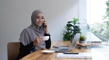 gelukkige mooie moslim vrouwelijke werknemer met hete koffiemok zittend op kantoor en het gebruik van mobiele telefoon kijken naar online nieuws ontspannen. foto