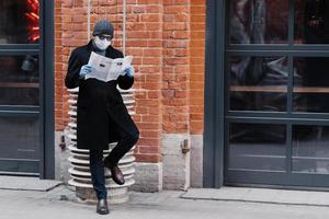 volledige opname van een serieuze man gekleed in een zwarte jas, draagt een zonnebril en een medisch masker, leest de krant, poseert tegen een bakstenen muur, voorkomt dat hij coronavirus krijgt. epidemie en quarantaine foto