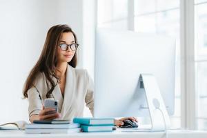 mooie vrouwelijke ondernemer met lang donker haar, werkt op de computer, ontwikkelt een reclamecampagnestrategie, poseert in een modern coworking-kantoor, gebruikt mobiele telefoon om online te chatten, draagt een bril foto