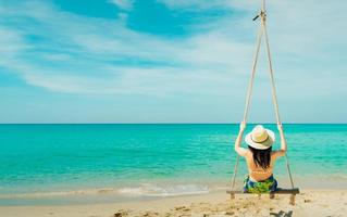 aziatische vrouw draagt badkleding en hoed zwaait met de schommels op het zandstrand en ziet er op zonnige dag prachtig uit in een tropisch paradijs, zee en lucht. zomervakantie. zomerse sferen. genietend en ontspannend meisje op vakantie foto