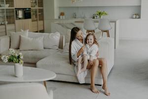 gelukkige schattige kleine handen poses op moeders benen gekleed in zachte witte badhanddoek voelt erg blij. moeder en dochter dragen huishoudelijke kleding en zitten op een comfortabele bank in de woonkamer. interieur foto