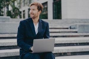 jonge glimlachende kantoormedewerker die buiten op de trap in het stadscentrum zit en een laptop op zijn schoot houdt foto