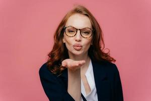 mooie vrouw met rood haar houdt lippen gevouwen, stuurt luchtkus, drukt liefde uit aan vriendje draagt optische bril, gekleed in zwart pak, houdt handpalm uitgestrekt bij mond, geïsoleerd op roze muur foto