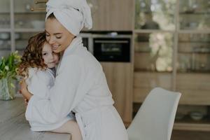 gelukkige aanhankelijke jonge moeder in badhanddoek en kamerjas omarmt met liefde die aan tafel zit, voelt zich verfrist na het douchen breng vrije tijd thuis door. familie schoonheid wellness concept foto