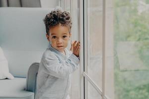 portret van schattig gemengd ras gekrulde babyjongen wachtend op ouders in de buurt van raam foto