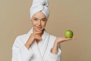 mooie gezonde vrouw raakt kin zachtjes aan houdt groene appel heeft een perfecte gladde huid draagt een zacht wit huisgewaad en gewikkelde handdoek op het hoofd modellen binnen met vers fruit. huid behandeling. foto