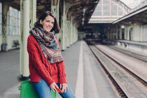 blije jonge europese vrouw draagt rode trui, sjaal, houdt mobiele telefoon vast, zit aan tas, geniet van reizen, poseert op treinstation, glimlacht vrolijk. mensen, toerisme, reizen en technologie concept foto
