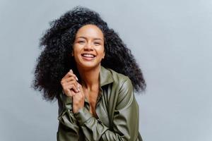 vrolijke vrouw met donkere huid, afro kapsel, houdt de handen bij elkaar, glimlacht oprecht naar de camera, toont witte tanden, draagt leren shirt, poseert over grijze achtergrond. mensen, emoties, etniciteit foto