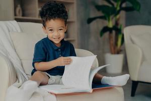 schattige kleine Afro-Amerikaanse jongen zittend in een fauteuil met verhalenboek foto