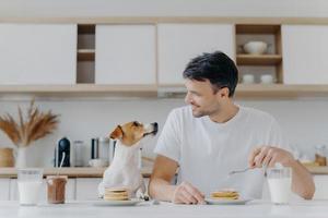 knappe brunet man kijkt blij naar zijn huisdier, heeft een zoet dessert als ontbijt, geniet van het weekend heeft een goede relatie met huisdier pose in keukeninterieur in modern appartement. mensen, voeding, dieren foto