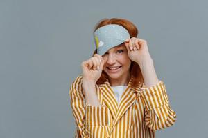 speelse vrouw met rood haar kijkt uit slaapmasker, draagt gestreepte pyjama's, heeft een vrolijke uitdrukking, poseert over grijze achtergrond, slaapt graag thuis. gezond slaap- en ontspanningsconcept foto