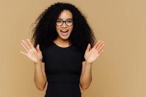 vrolijke speelse afro-amerikaanse vrouw heft handpalmen op, voelt zich opgewonden, drukt geluk uit, blij om cadeau te ontvangen, draagt casual outfit, bril, staat tegen bruine achtergrond. positieve reactie foto