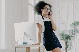 tevreden vrouwelijke kantoormedewerker met krullen gekleed in zwarte sarafan, leunt aan tafel, poseert in kast bij computermonitor, heeft telefoongesprek, bespreekt details van contract, heeft een glimlach op het gezicht foto