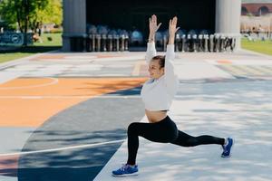 flexibele pasvorm sportvrouw doet sportoefeningen buitenshuis houdt de armen omhoog heeft een blije uitdrukking draagt hoodies leggings en trainers poses op stadion in stedelijke plaats luistert naar favoriete audiotrack. foto