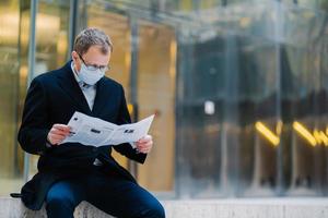 buitenopname van serieuze man baas neemt pauze na wandelen, leest krant, draagt bril voor goed zicht, medisch masker om zichzelf te beschermen tegen virussen, ontdekt nieuws over verspreiding coronavirus foto