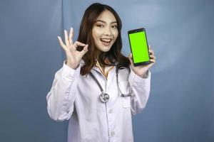 jonge aziatische vrouwelijke arts wijst en toont groen scherm of kopieert ruimte op haar smartphone. duimen opdagen of ok teken. foto