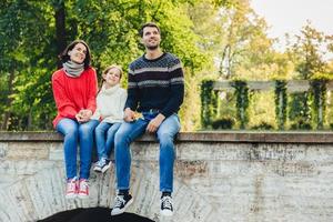 horizontaal portret van schattig kind zit tussen haar moeder en vader, geniet van prachtige landschappen vanaf de top van een oude brug, adem frisse lucht in, kijk in de verte. familie en relatie concept foto